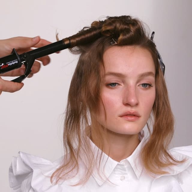 Kvinne med brunt hår med hårklyper i og en hånd som krøller håret hennes med en BaByliss krølltang