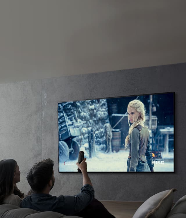 Par som sitter i en soffa och tittar på en film där det står Netflix i ena hörnet med en ljus kvinna i gammeldags kläder