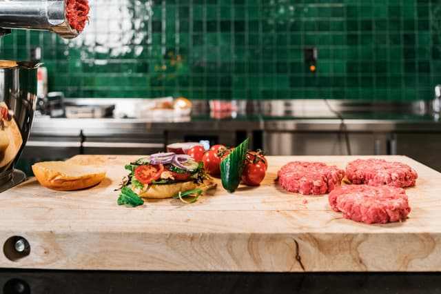 Hamburgerbröd med grönsaker och tre råa burgare vid sidan på en skärbräda av trä och kött kommer ut ur en köttkvarn