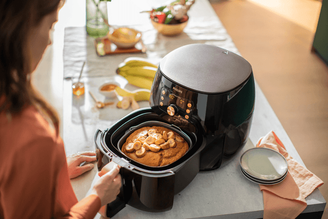 Kvinna placerar mat i en airfryer på ett bord eller en köksö med frukt och assietter bredvid.