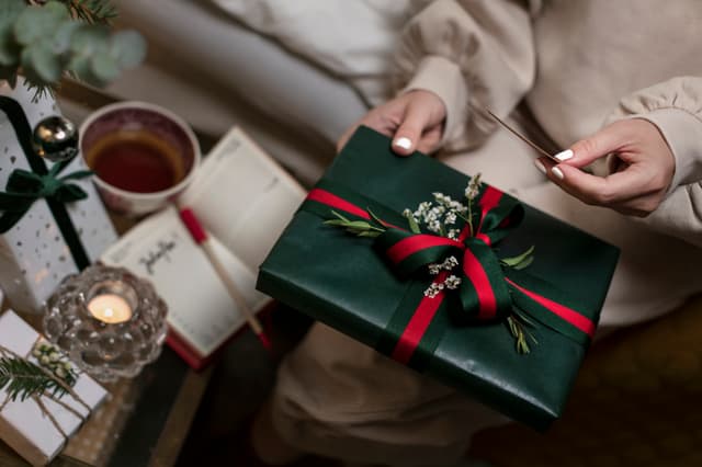 En kvinna med vitt nagellack och julklappar och en kaffekopp på ett bord bredvid sig håller en julklapp och ett kort i handen 