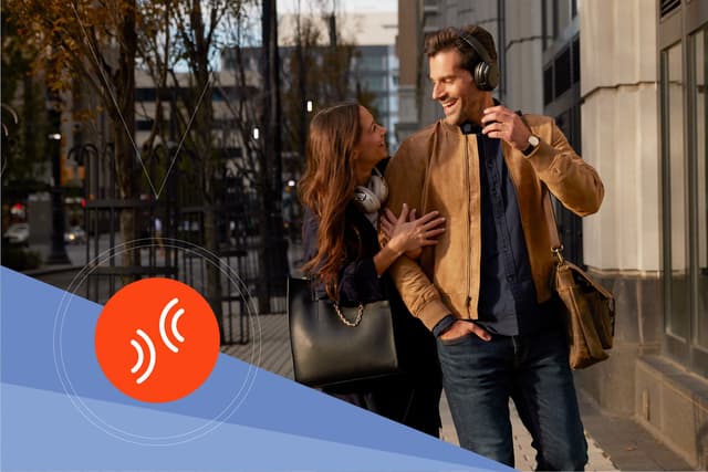 JBLs logo til Smart Ambient-teknologien og JBL Tour One M2 høretelefoner på både en mand og en kvinde, der går tur sammen