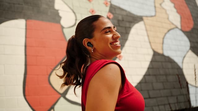 Hymyilevä tyttö Bose QuietComfort Ultra Earbuds -nappikuulokkeet päässään ja värikäs seinä taustalla