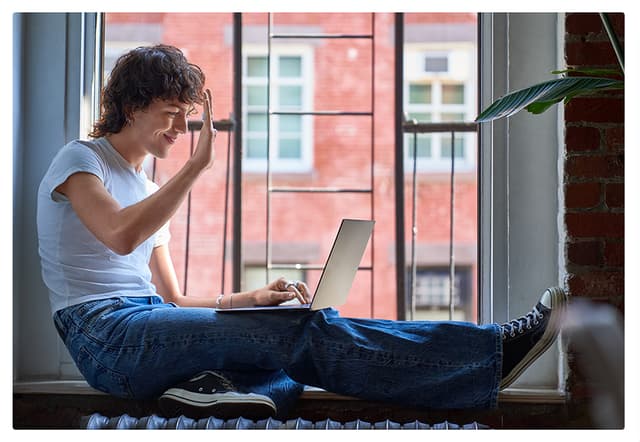 Person sitter i vinduskarm med laptop på fanget og vinker inn i kameraet