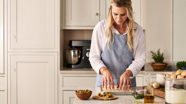 En kvinne baker ut en focaccia-deig på en kjøkkenøy.