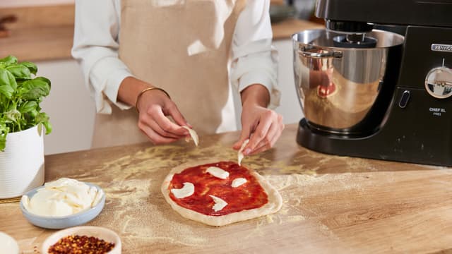 En kvinne tilbereder hjemmelaget pizza på kjøkkenbenk ved siden av en svart Kenwood Chef XL Pro-kjøkkenmaskin.