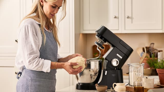 En kvinne står foran en svart Kenwood Chef XL Pro-kjøkkenmaskin med en deig i hendene.