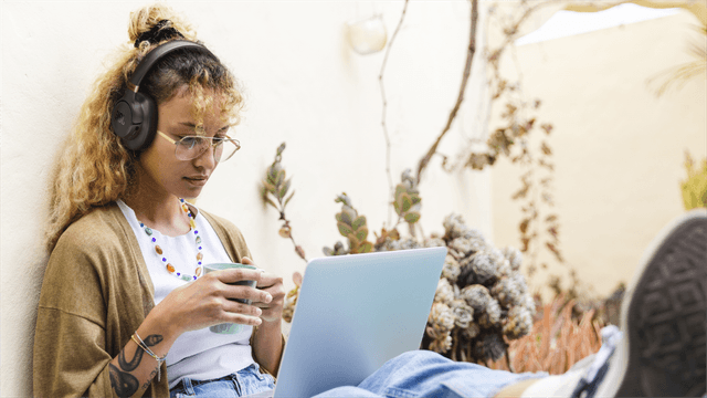 En kvinne sitter mot en murvegg med JBL Tune 785-hodetelefoner på og en laptop på fanget.