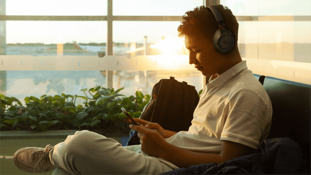 En mann sitter i en flyplasslounge og ser på mobilen med JBL Tune 785-hodetelefoner på.