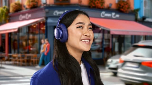 En kvinne står i en travel handlegate med mørkeblå JBL Live 780 støydempende hodetelefoner på.