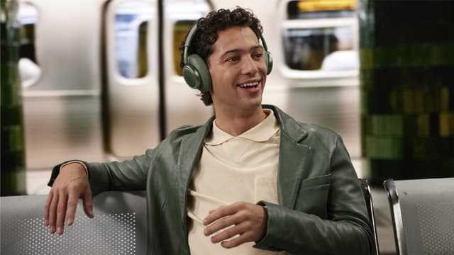 Mies istuu juna-asemalla ja puhuu puhelimeen valkoiset JBL Live 780 NC -kuulokkeet päässään