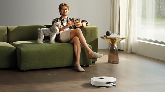 En mann og en hund sitter på en grønn sofa i en stue mens robotstøvsugeren Dreame L50 Ultra AE vasker gulvet rundt dem.