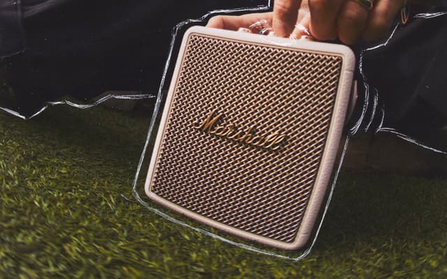 Closeup of a beige Marshall Stockwell III speaker outdoors being carried over a lawn.