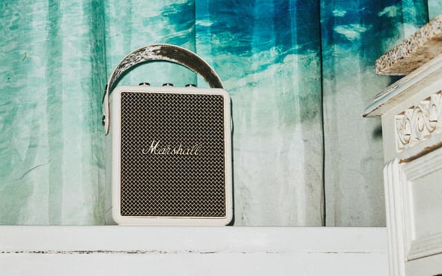 A beige Marshall Stockwell III speaker on a white furniture in front of a turquoise curtain.
