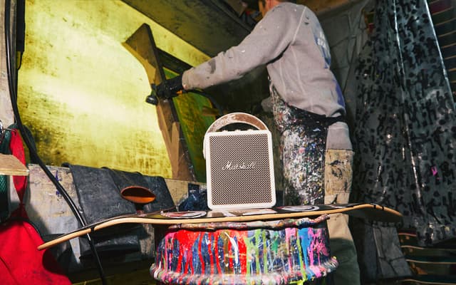 A man working on a wall next to his skateboard and a beige Marshall Stockwell III speaker.