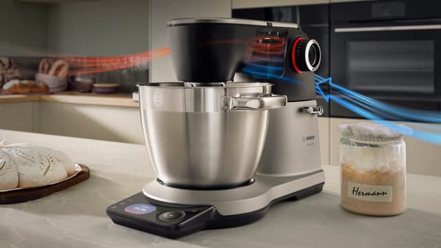En Bosch Serie 8-kjøkkenmaskin på en kjøkkenbenk mellom et brød i en brødform og en surdeigsstarter på glass.