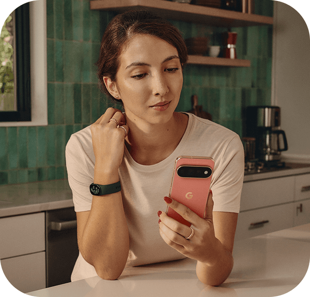 En kvinde i et køkken med et Fitbit Edge på håndleddet, der kigger på sin Google Pixel smartphone