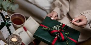 En kvinna med vitt nagellack och julklappar och en kaffekopp på ett bord bredvid sig håller en julklapp och ett kort i handen 