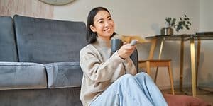 Hymyilevä nainen istuu lattialla sohvaan nojaten ja osoittaa televisiota kaukosäätimellä