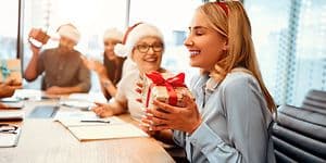 Glad kvinde som holder en julegave imens hun sidder omkring sine kollegaer på kontoret