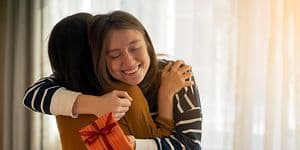 Hymyilevä nainen halaa toista naista samalla kun pitelee joululahjapakettia kädessään