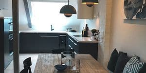 Köket sett från matbordet i trä, stilren design med grå färg och vit bänkskiva anpassat till lutande väggar och takbjälkar. . 