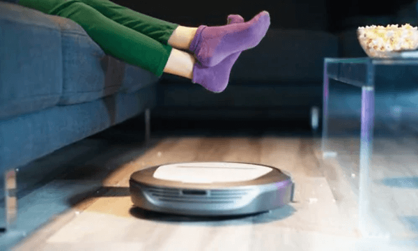Robotdamsugare städar på golvet medan en person med lila strumpor slappar i soffan. En popcorn skål står på soffbordet. 
