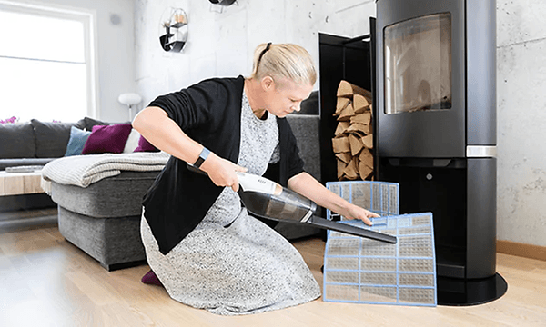 Kvinna sitter böjd på golvet framför en öppen spis med en dammsugare i högsta hugg och städar av filtret till värmepumpen. 