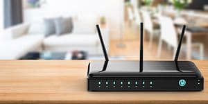 En svart trådlös router med tre antenner, i bakgrunden i oskärpa syns en soffa, TV och ett stort matbord. 