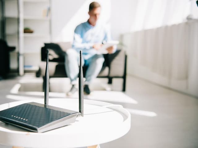 Trådlös router placerad på ett vitt bord, i bakgrunden i oskärpa syns en man som sitter i en fåtölj.