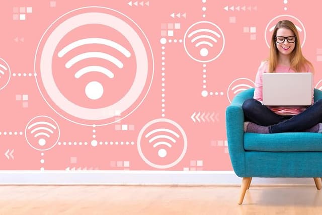 Kvinna sitter med korslagda ben i en turkos fåtölj oc har en laptop i knät. Väggen bakom är rosa med en massa WiFi-symboler.