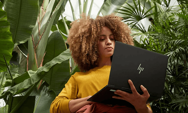 Kvinde står under nogle tropiske planter og holder en HP Envy PC