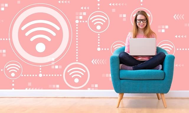 Kvinde sider i en turkisblå lænestol og arbejder på en bærbar. Bag hende er den lyserøde væg dækket af wi-fi-symboler