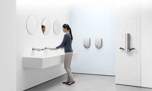 Dyson Airblade håndtørkere montert på vegg på hvitt bad hvor en kvinne vasker hendene