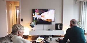 Ældre kvinde sidder i en sofa med hendes tablet, mens hendes mand sidder og ser TV