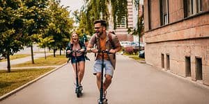 Man och kvinna i full fart på en väg på varsin elsparkcykel. 
