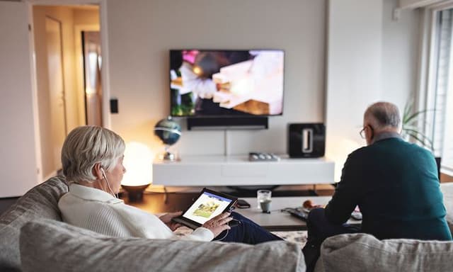 Eldre kvinne som sitter i sofa med nettbrett, mens eldre mann ser på TV i bakgrunnen