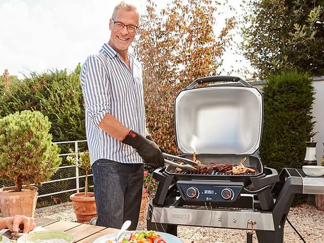 En man grillar på en elektrisk grill på en uteplats och tittar på en kvinna som sitter vid ett bord och ler mot mannen.