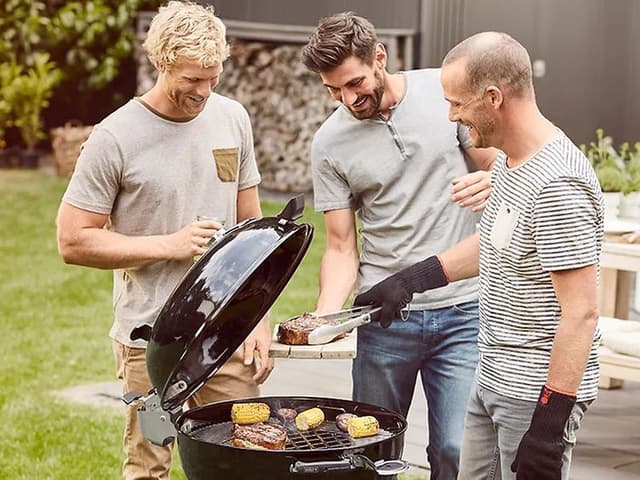 Tre män har samlats runt en klassisk rund svart kolgrill från Weber på en uteplats i en trädgård.