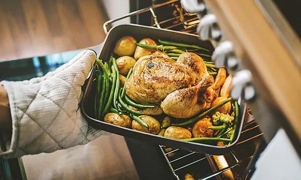 En hand i grytvante tar ut en form med tillagad kycklingrätt med potatis från ugnen. 