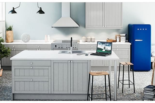 Ljust kök med en vit väggmonterad köksfläkt och ett blått kylskåp från Smeg och köksö med stolar. 
