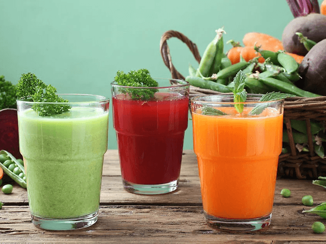 Tre glas med juice på ett bord med grönsaker. En grön juice, en orange och en röd.