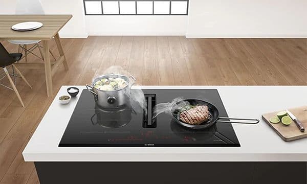 Bosch induktionshäll med inbyggd frånluftsfläkt på en köksö. På spisen står kastrull med grönskaer och stekpanna med köttbit. 