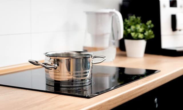 En stor kastrull står på en platta på en induktionshäll, i bakgrunden syns en vit vattenkokare och en krukväxt. 