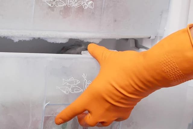 Hand i orange diskhandske öppnar en igenfrusen fryslåda i en frys som behöver frostas av. 