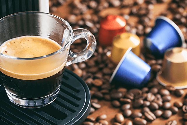En kopp espresso fra en kapselmaskin med kapsler og kaffebønner på kjøkkenbenk