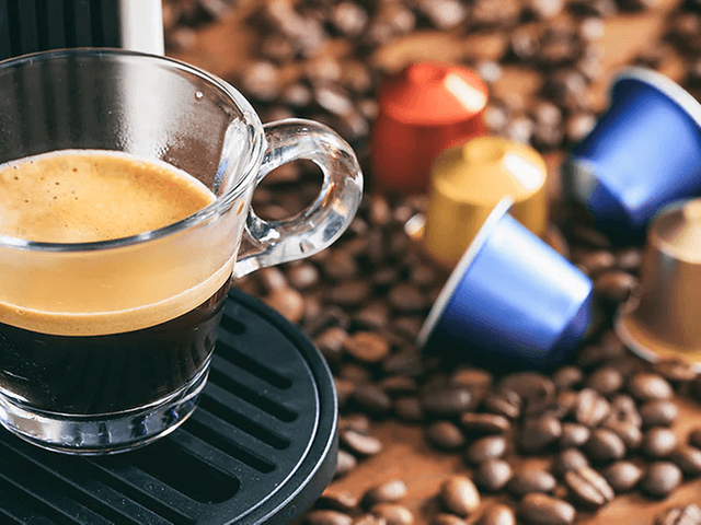 En kopp espresso fra en kapselmaskin med kapsler og kaffebønner på kjøkkenbenk