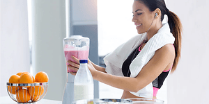 Urheilullisiin vaatteisiin pukeutunut nainen tekee vaaleanpunaista smoothieta tehosekoittimella