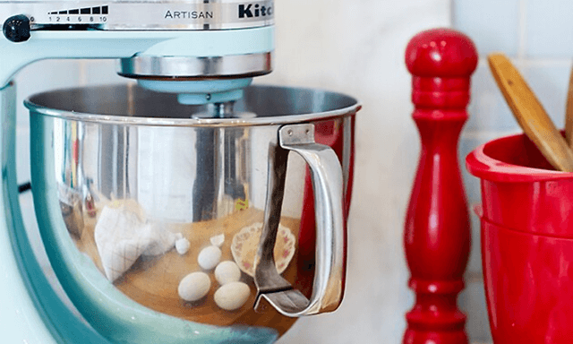 en blå KitchenAid-kjøkkenmaskin på et bord ved siden av egg og rødt kjøkkenutstyr