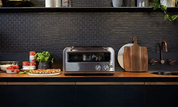 Sage-pizzauuni keittiön työtasolla, jonka vieressä tuore pizza ja tomaatteja